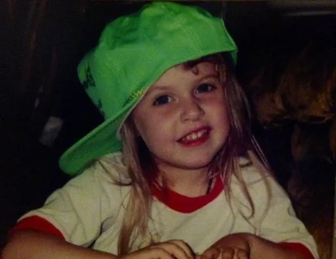A young girl with light brown hair wearing a green cap and a white and red shirt, smiling at the camera.