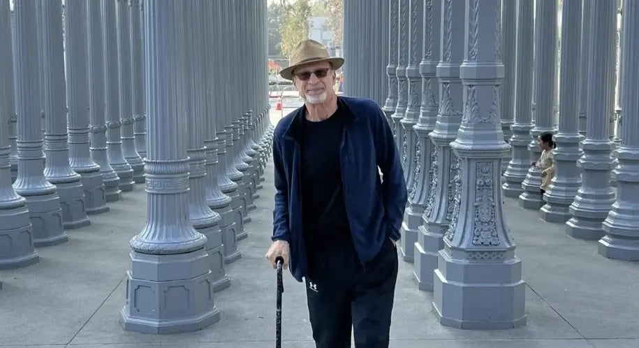 Man walking with a cane through an outdoor art installation of tall lamp posts, smiling during his recovery