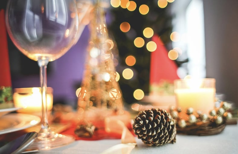 Close-up of a festive holiday table setting with candles, pinecones, and wine glasses, with blurred Christmas lights in the background