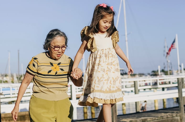 Elderly woman holding hands with a young girl as they walk along a marina dock on a sunny day