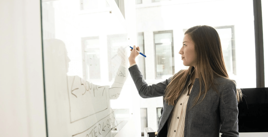 A woman writing or drawing on a transparent whiteboard in an office setting.