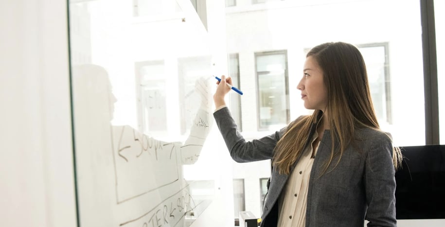 A woman writing or drawing on a transparent whiteboard in an office setting.