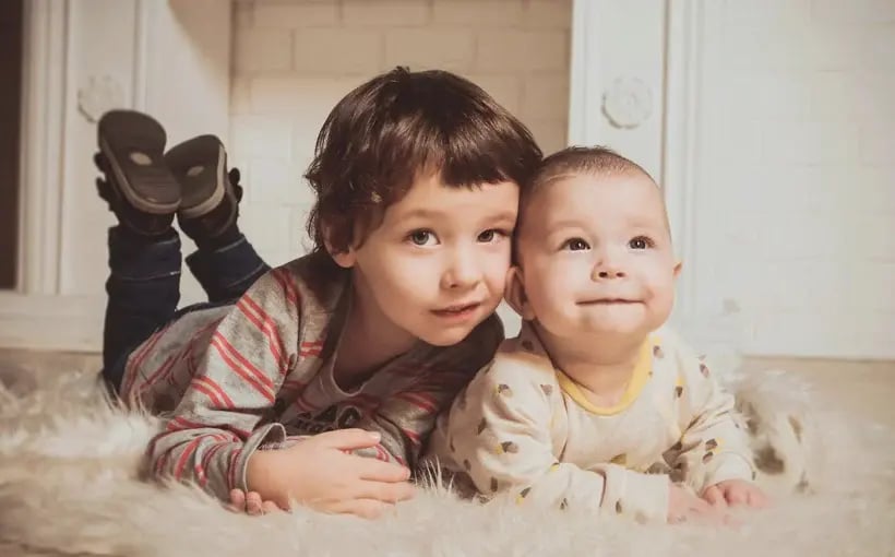 Two young siblings lying on a fluffy rug indoors, smiling and enjoying a cozy moment together