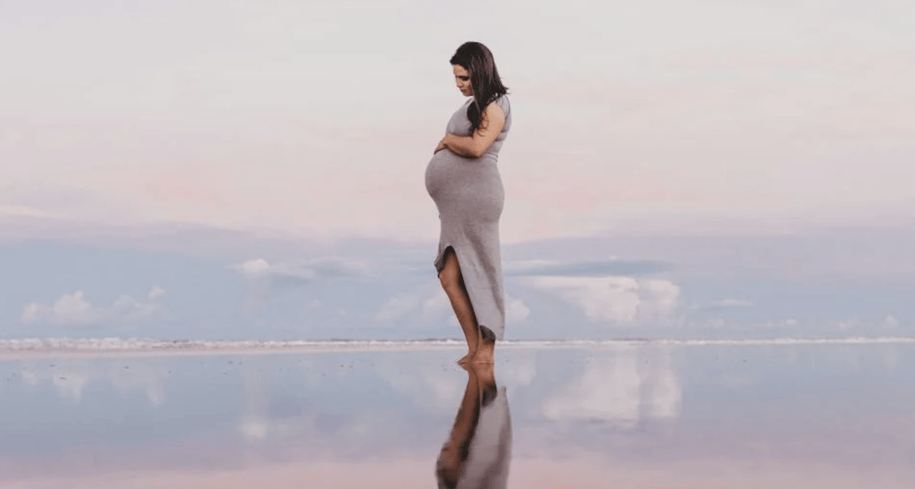 A pregnant woman in a tight dress standing on a beach.