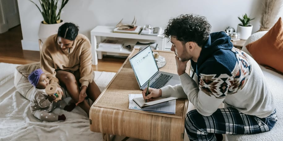 Two adults and a baby in a living room, with one adult working on a laptop.