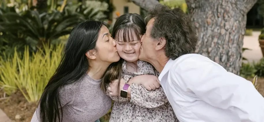 Young girl with Down syndrome smiling as she is kissed on both cheeks by two adults, capturing a moment of love and inclusion