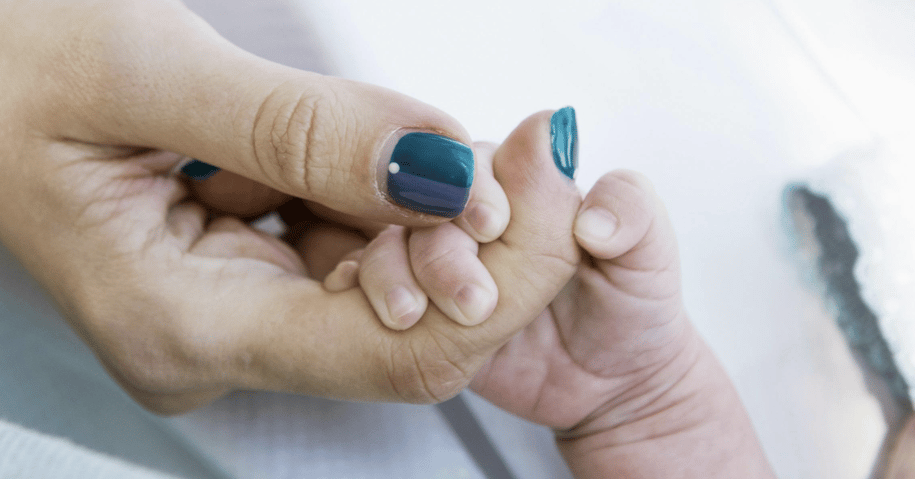 A close-up shot of an adult hand with dark nail polish gently holding the tiny hand of a newborn baby. The baby's fingers are curled around the adult's index finger.