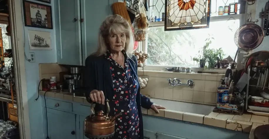 Elderly woman standing in a cozy kitchen holding a kettle, surrounded by vintage decor and household items