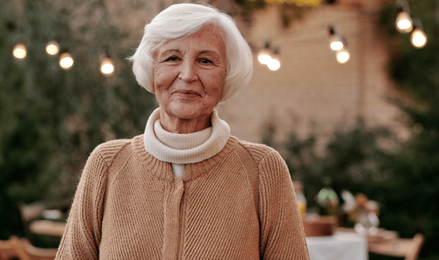 Elderly woman standing outdoors in a cozy sweater, looking content and peaceful with lights in the background