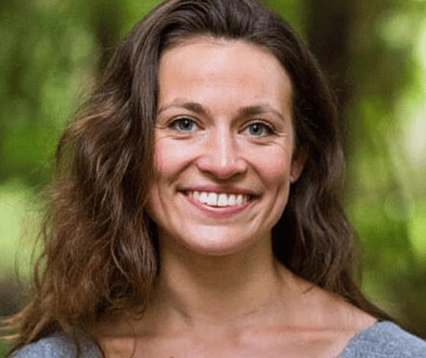A smiling woman with brown, wavy hair looking directly at the camera, outdoors.