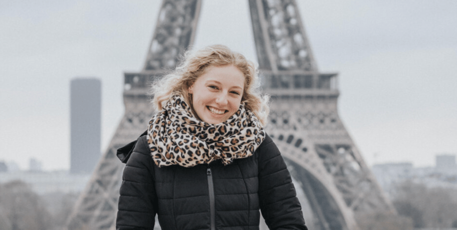 A smiling woman with blonde hair and a leopard print scarf stands in front of the Eiffel Tower.