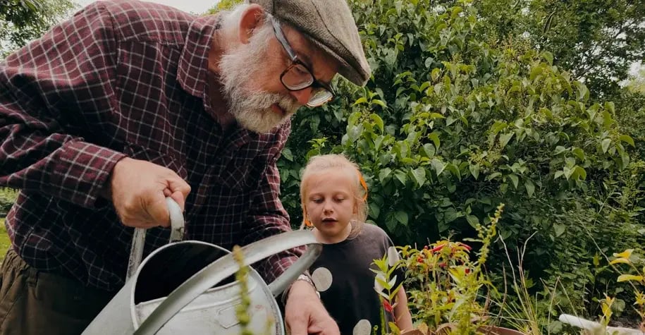 Older man watering plants with a young child in a garden, sharing a moment of intergenerational bonding