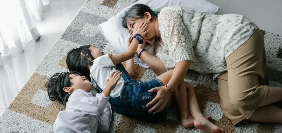 An adult, possibly a caregiver, is lying on the floor on a rug, interacting with two children. One child is lying on their stomach, and the other is lying on their back. The adult is smiling and appears to be playing with the children.