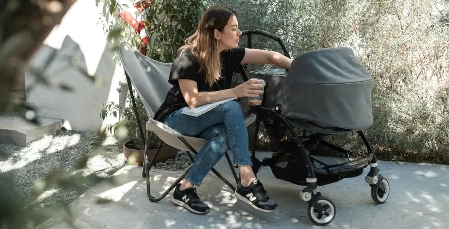 Woman sitting on a bench outdoors next to a baby stroller, sipping coffee and looking thoughtfully ahead