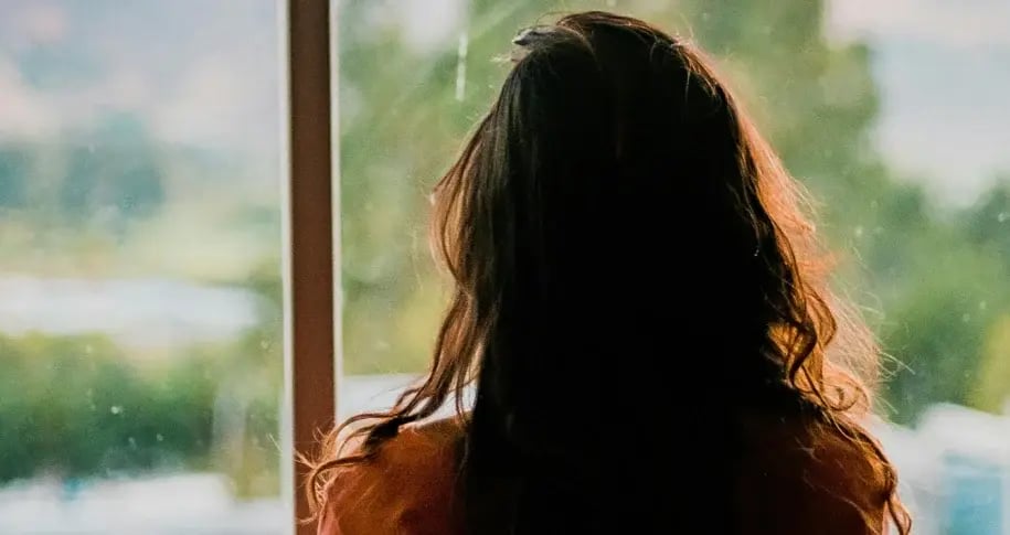 Person with long hair sitting alone and looking out a window at a blurred green landscape, conveying reflection and solitude