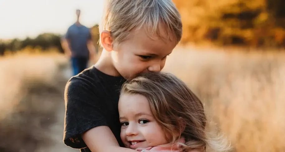 A young boy hugging a younger girl outdoors with a blurred background.