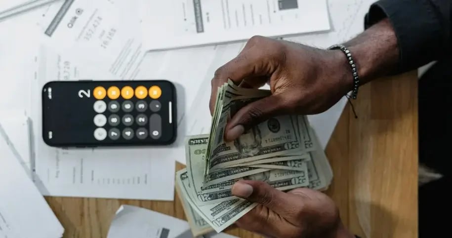 A person's hands counting a stack of US dollar bills next to a calculator.
