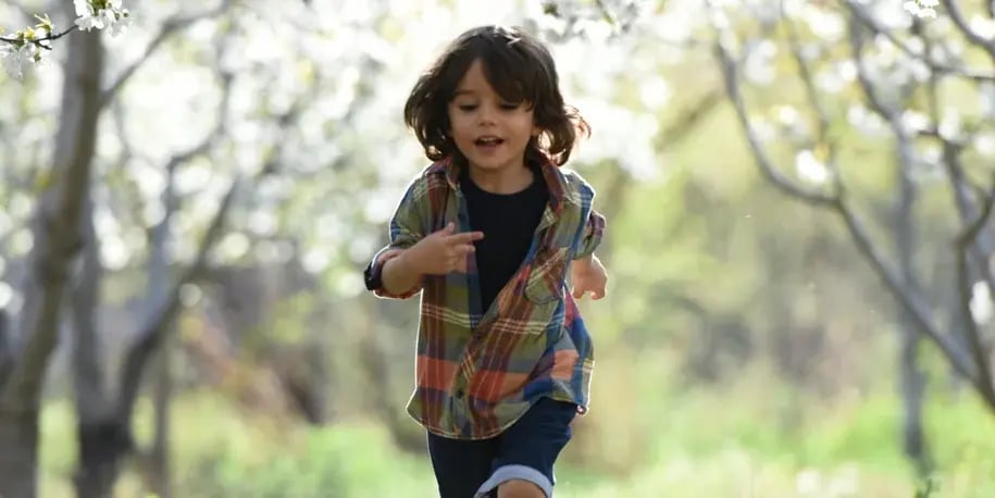 A young child with brown hair runs joyfully through a sunlit, leafy outdoor environment.