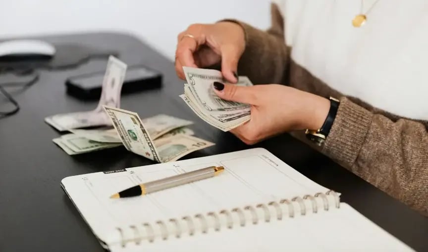 Close-up of hands counting dollar bills on a desk next to a notebook.