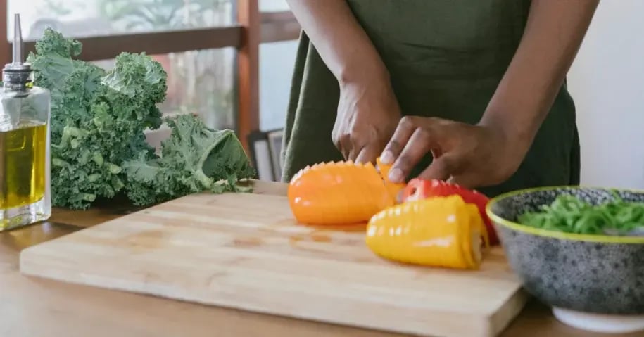 Hands chopping yellow and orange bell peppers on a wooden cutting board with leafy greens and olive oil nearby
