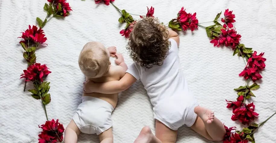 Two young siblings lying on a white blanket decorated with red flowers, sharing a tender moment together