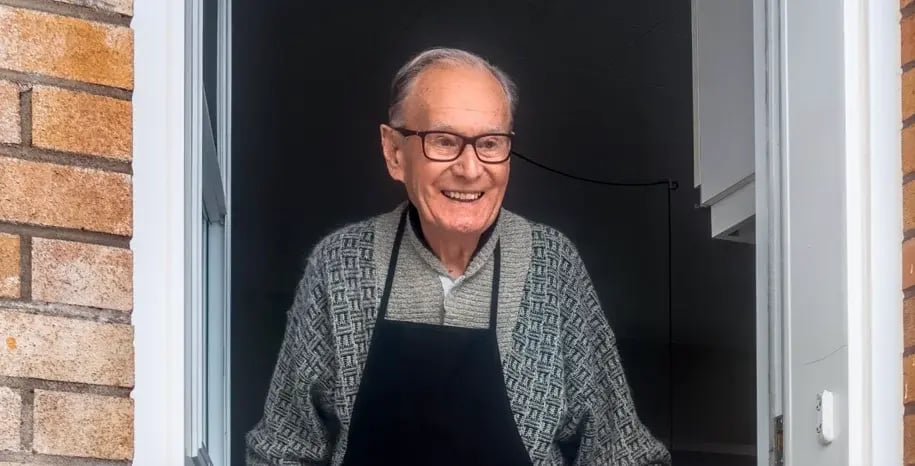 Smiling elderly man standing in a doorway wearing glasses and a dark apron