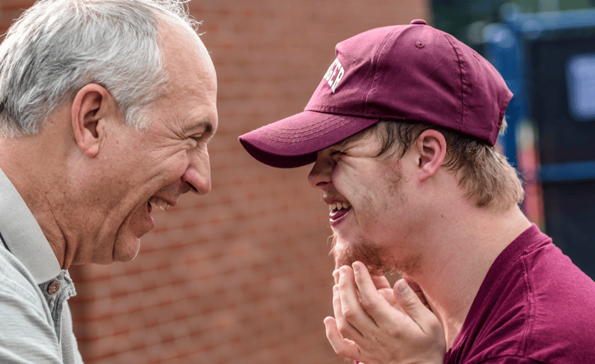 Older man and young adult with a disability sharing a joyful moment, smiling and leaning towards each other