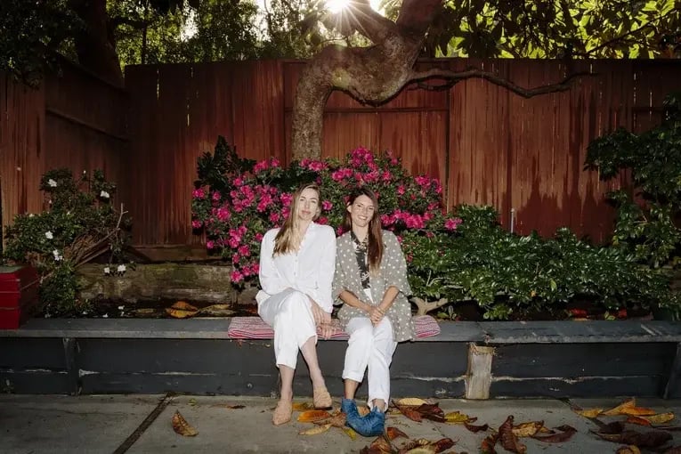 Two women sitting together on a bench surrounded by flowers, smiling and enjoying a calm outdoor moment